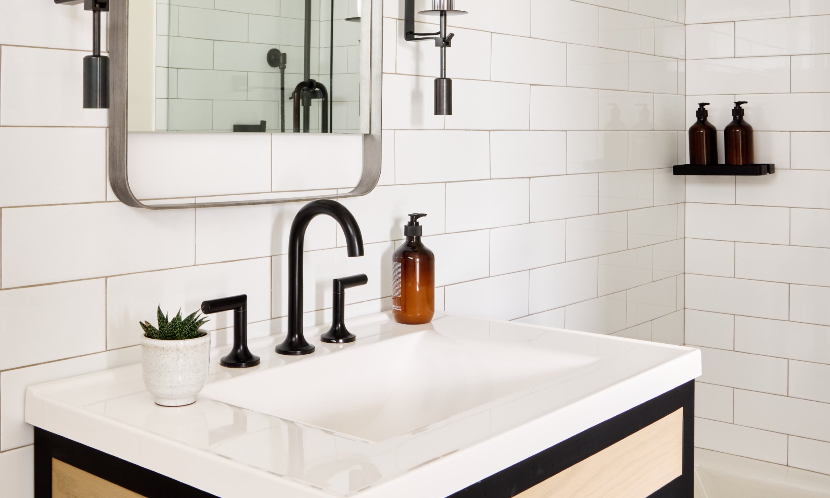 Modern bathroom with white subway tiles, black faucet, and framed mirror. Small potted plant and brown soap dispenser on the sink.