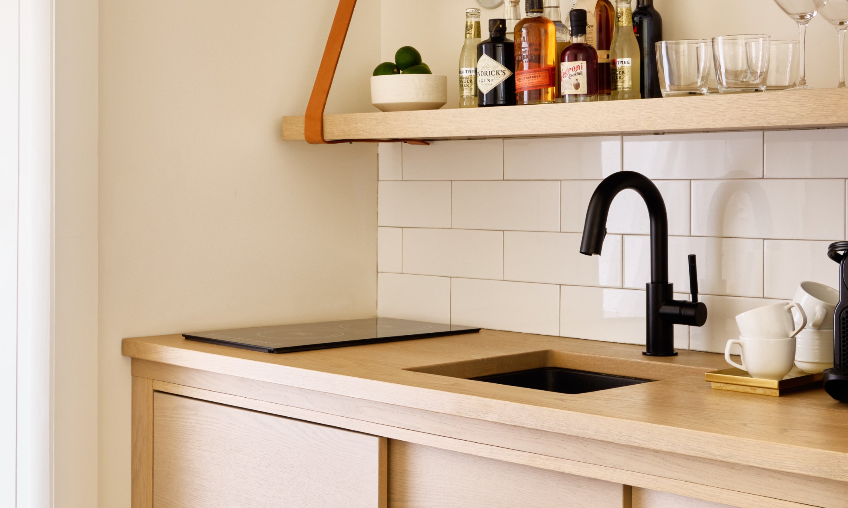Modern kitchen corner with wood countertop, black faucet, and assorted liquor bottles on a shelf. White tiled backsplash adds a clean look.