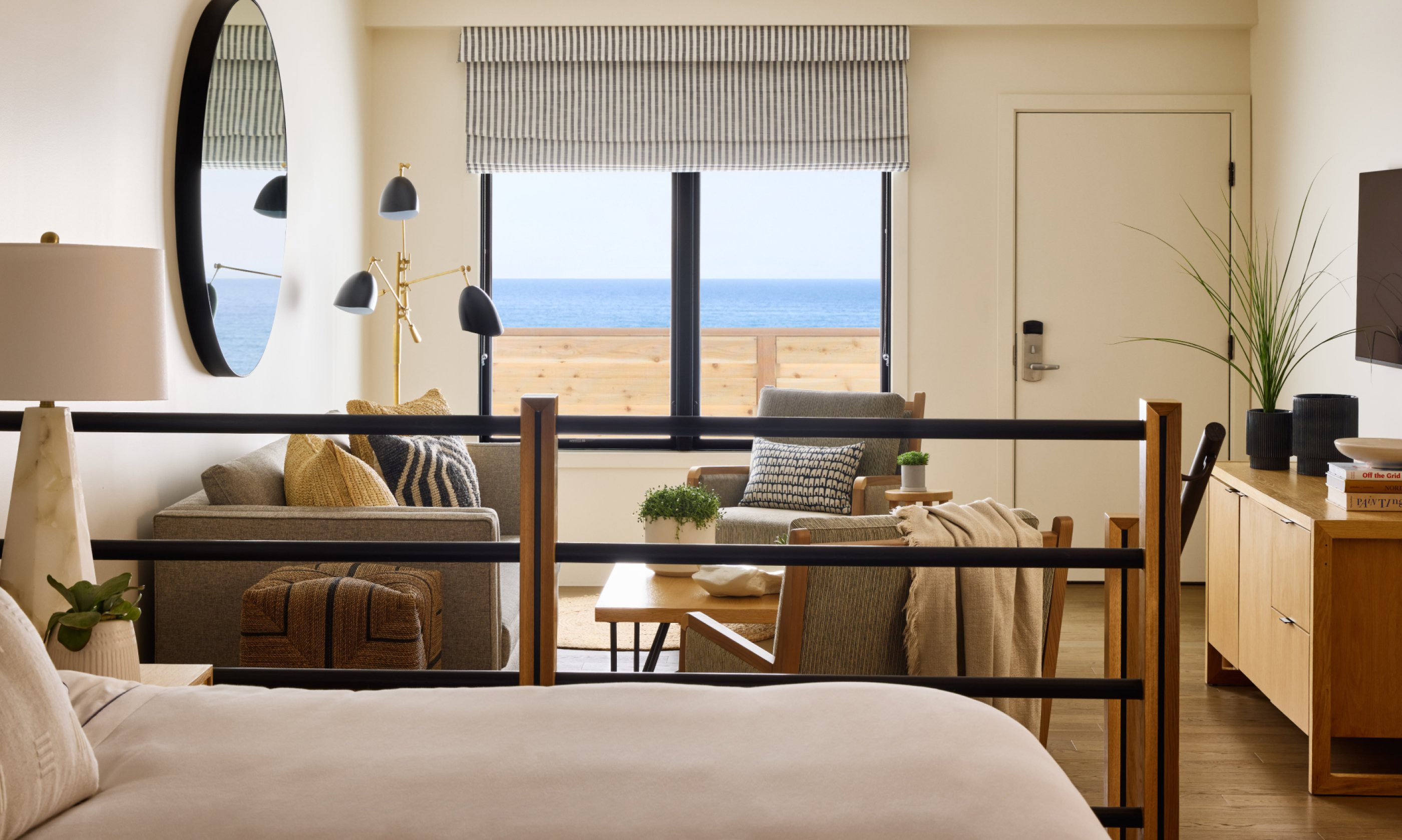 Modern hotel room with a bed, seating area, and ocean view window. Decor includes a large round mirror, stylish lamps, and a flatscreen TV.