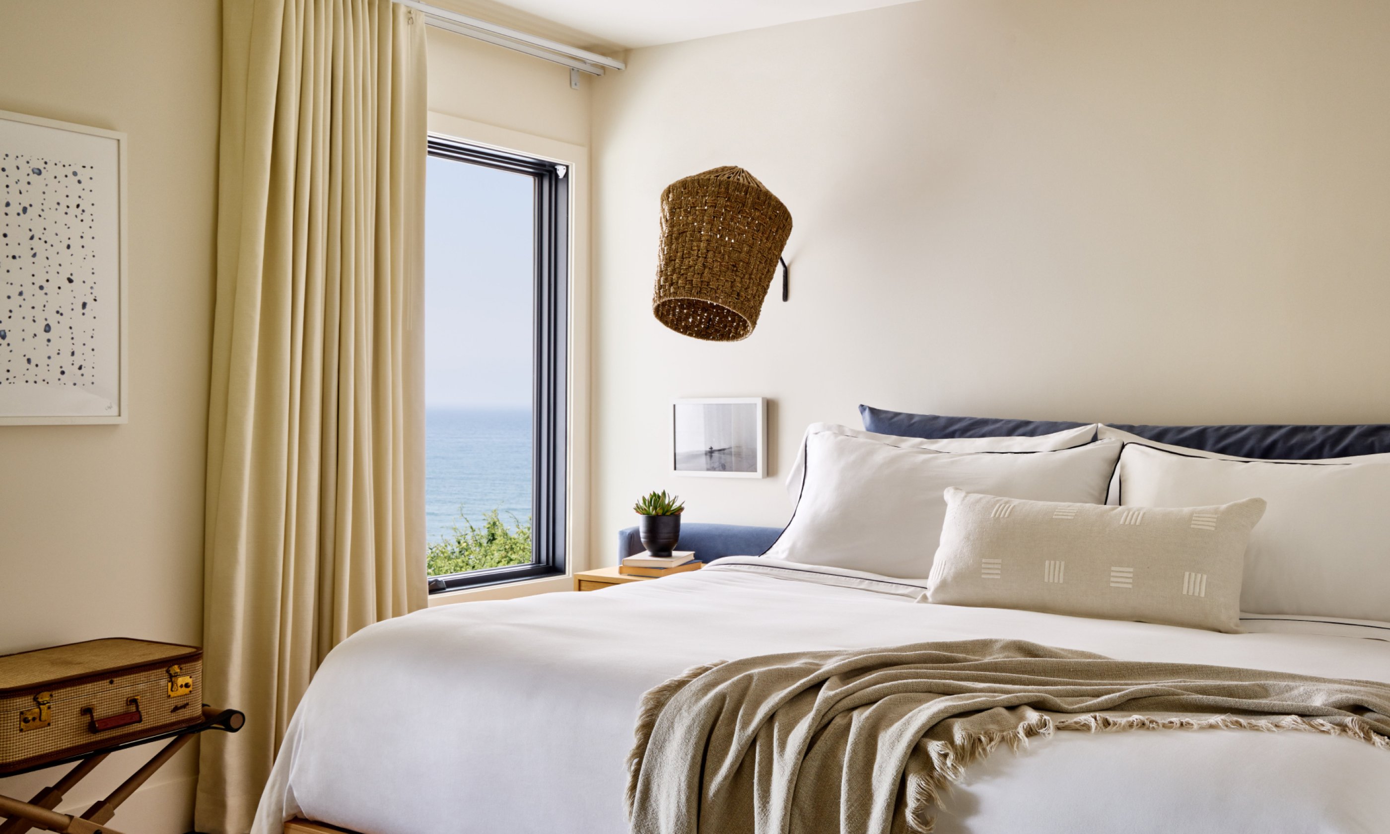 Cozy bedroom with a large bed, soft pillows, and a throw blanket. Ocean view through window, beige curtains, wall decorations, and potted plant.