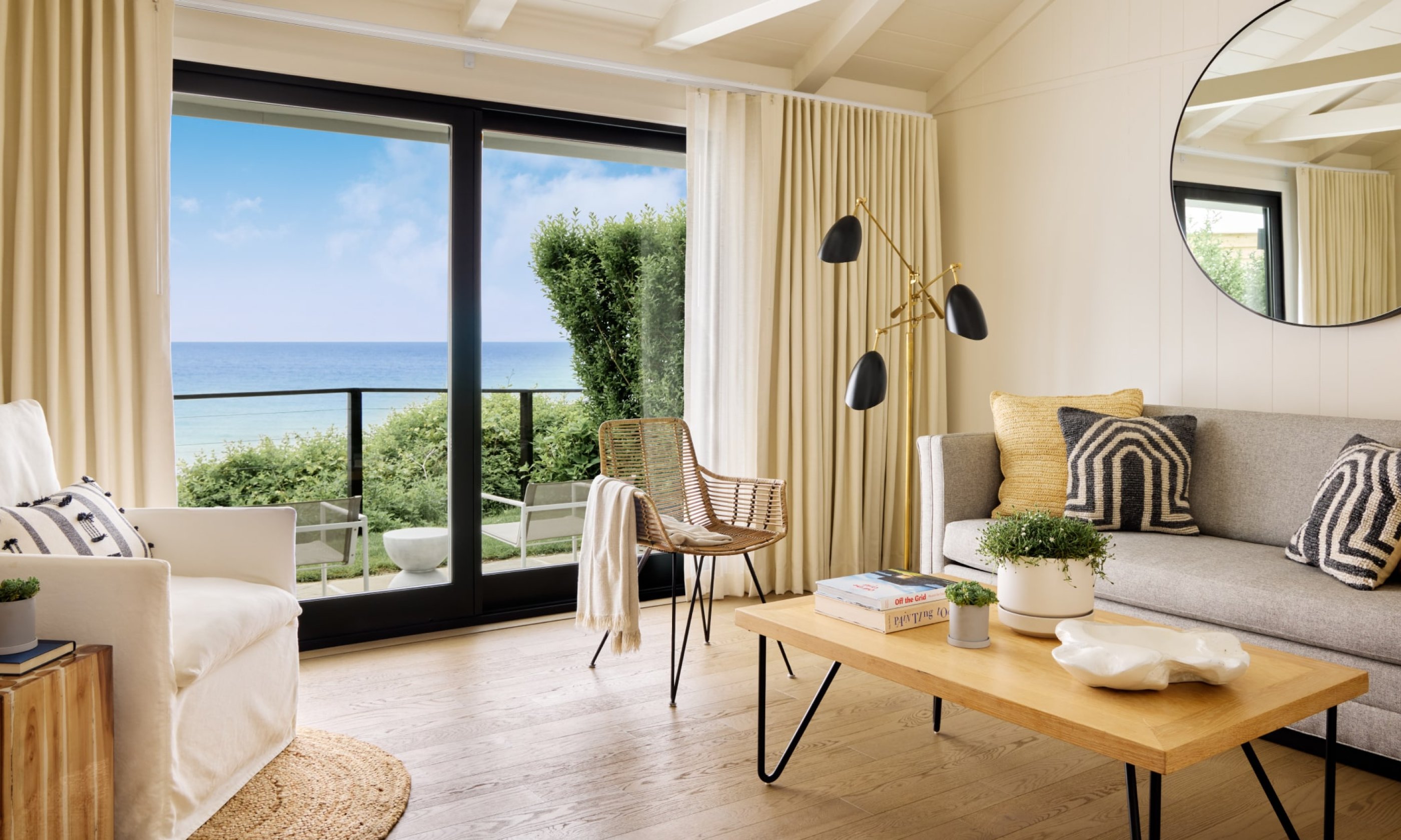 Cozy living room with beach view. Neutral tones, large window, gray sofa, patterned pillows, wooden coffee table, and modern lamp.