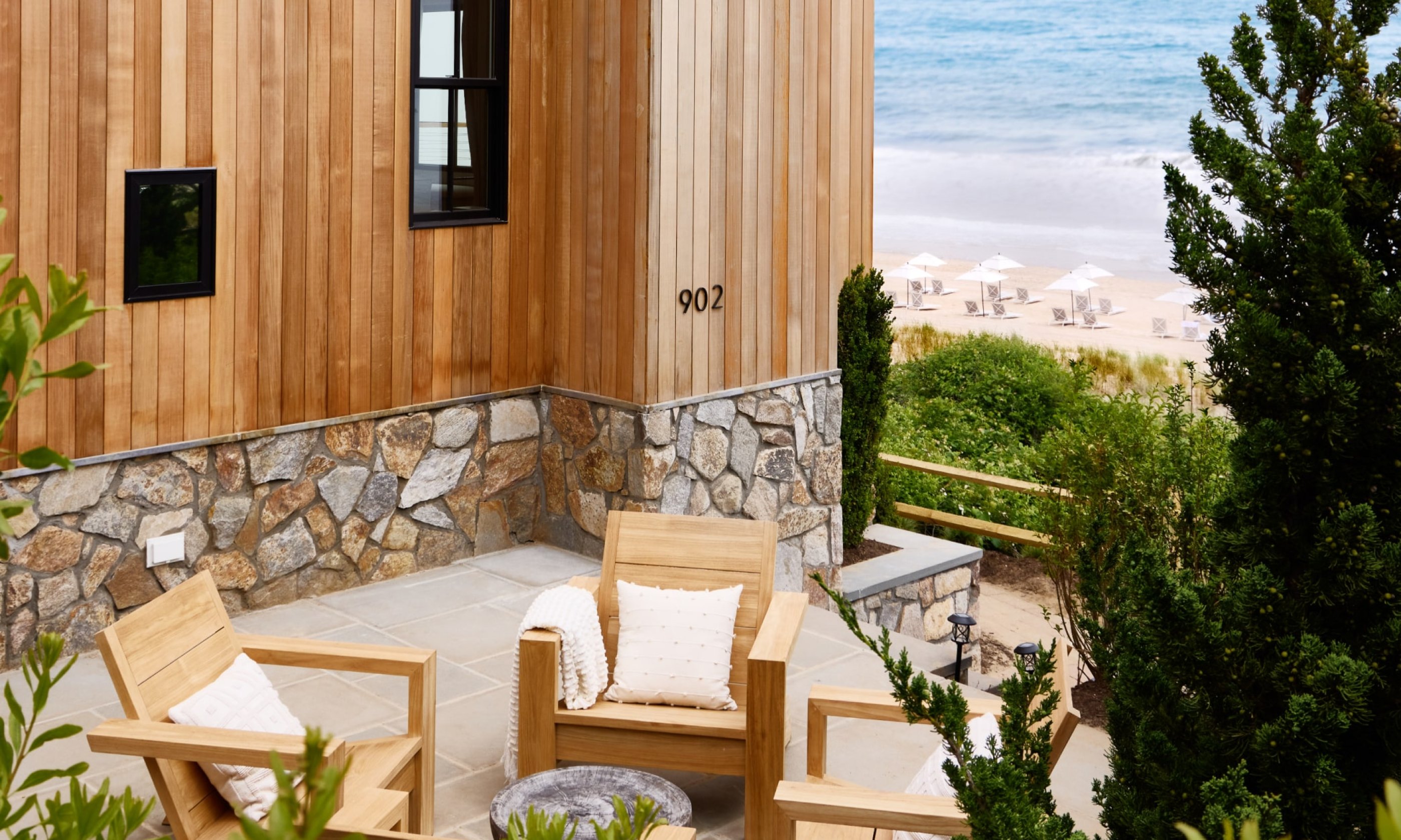 Wooden patio with cushioned chairs, overlooking a beach with gentle waves. Stone accents on the building, surrounded by greenery.