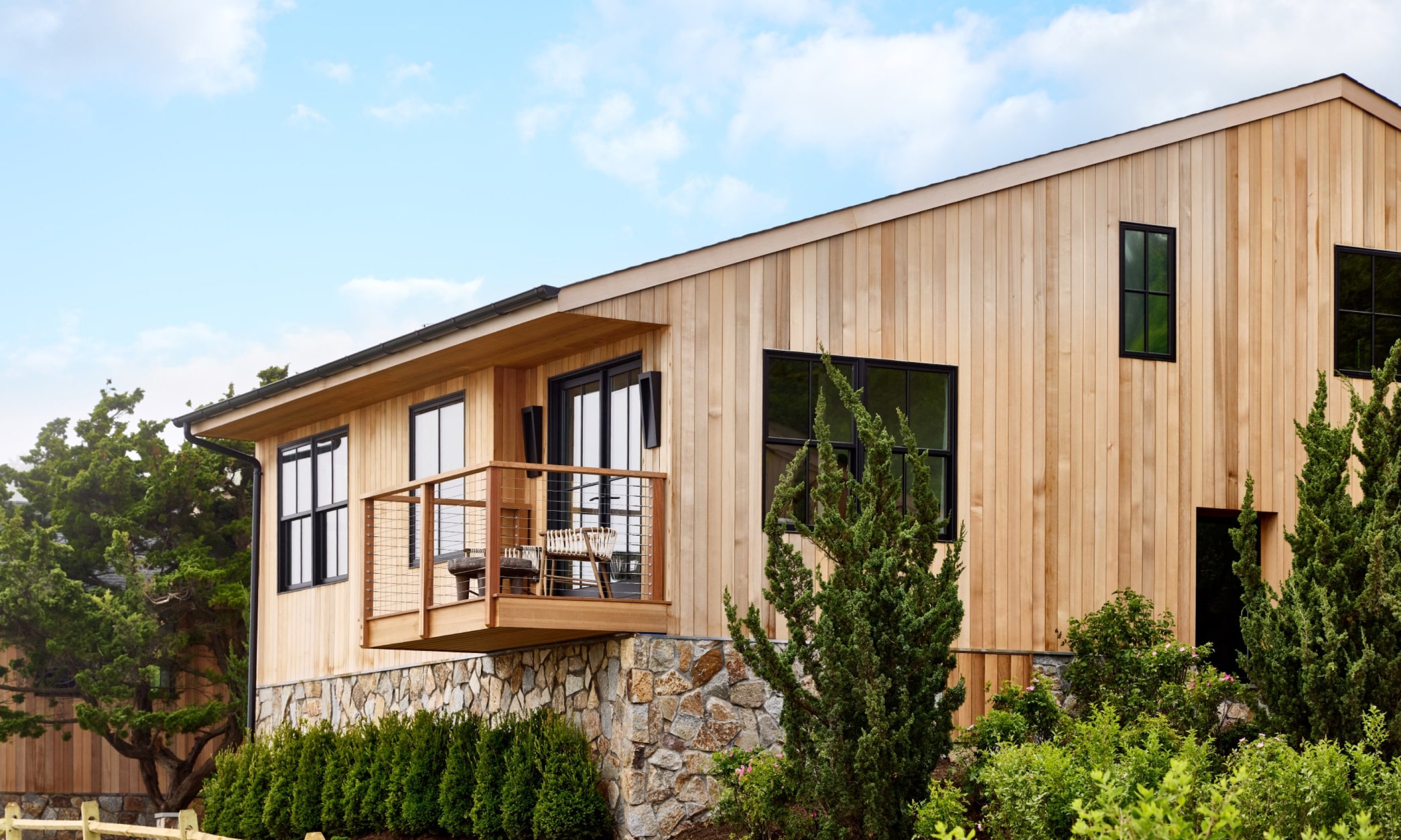 Modern wooden house with large windows, small balcony, and lush greenery. Blue sky with clouds in the background.
