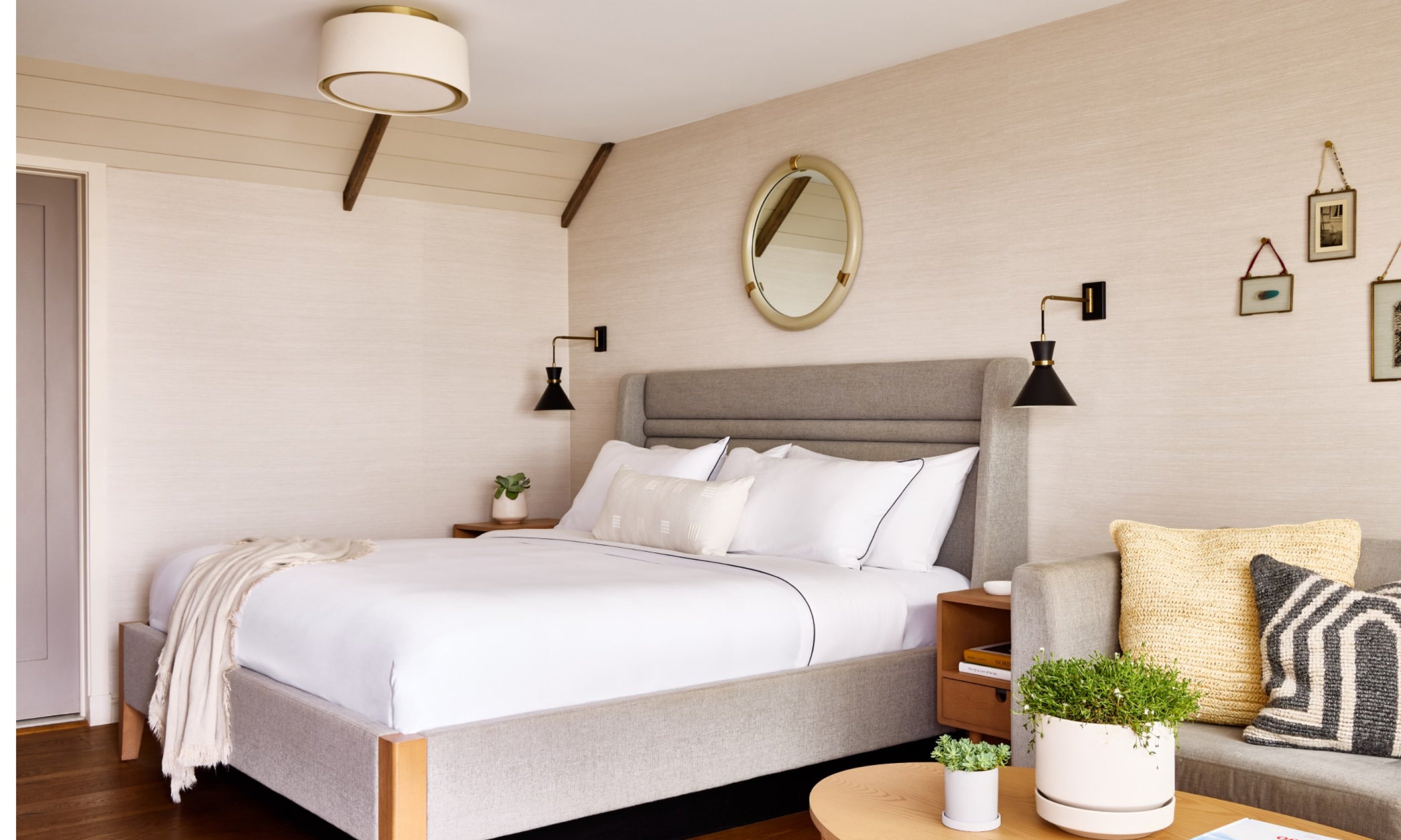 Bright bedroom with a large bed, round mirror, wall lamps, and cozy sofa. Neutral tones and small plants create a relaxing atmosphere.