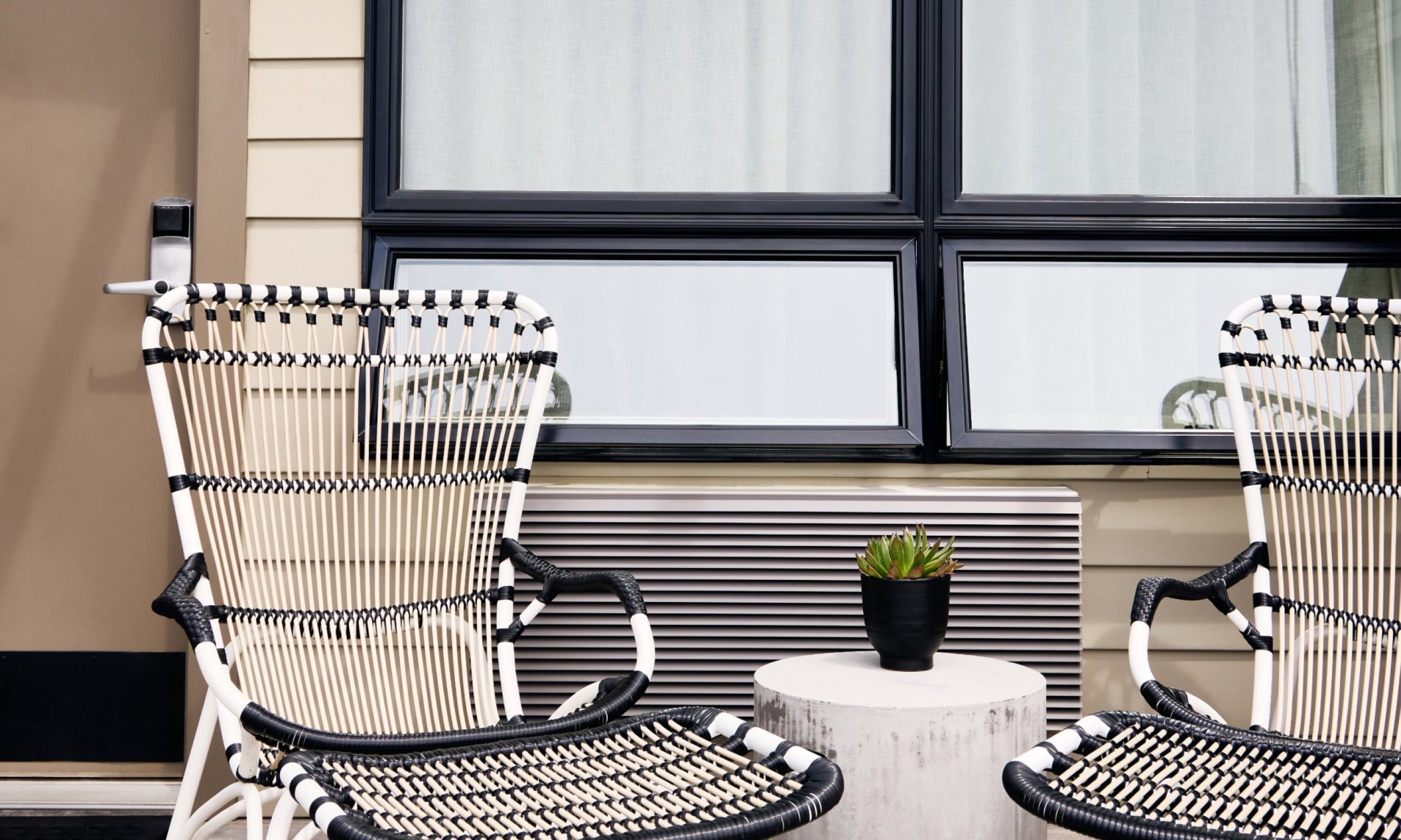 Black and white chairs on a patio facing a small table with a potted plant, set against a large window with white curtains.