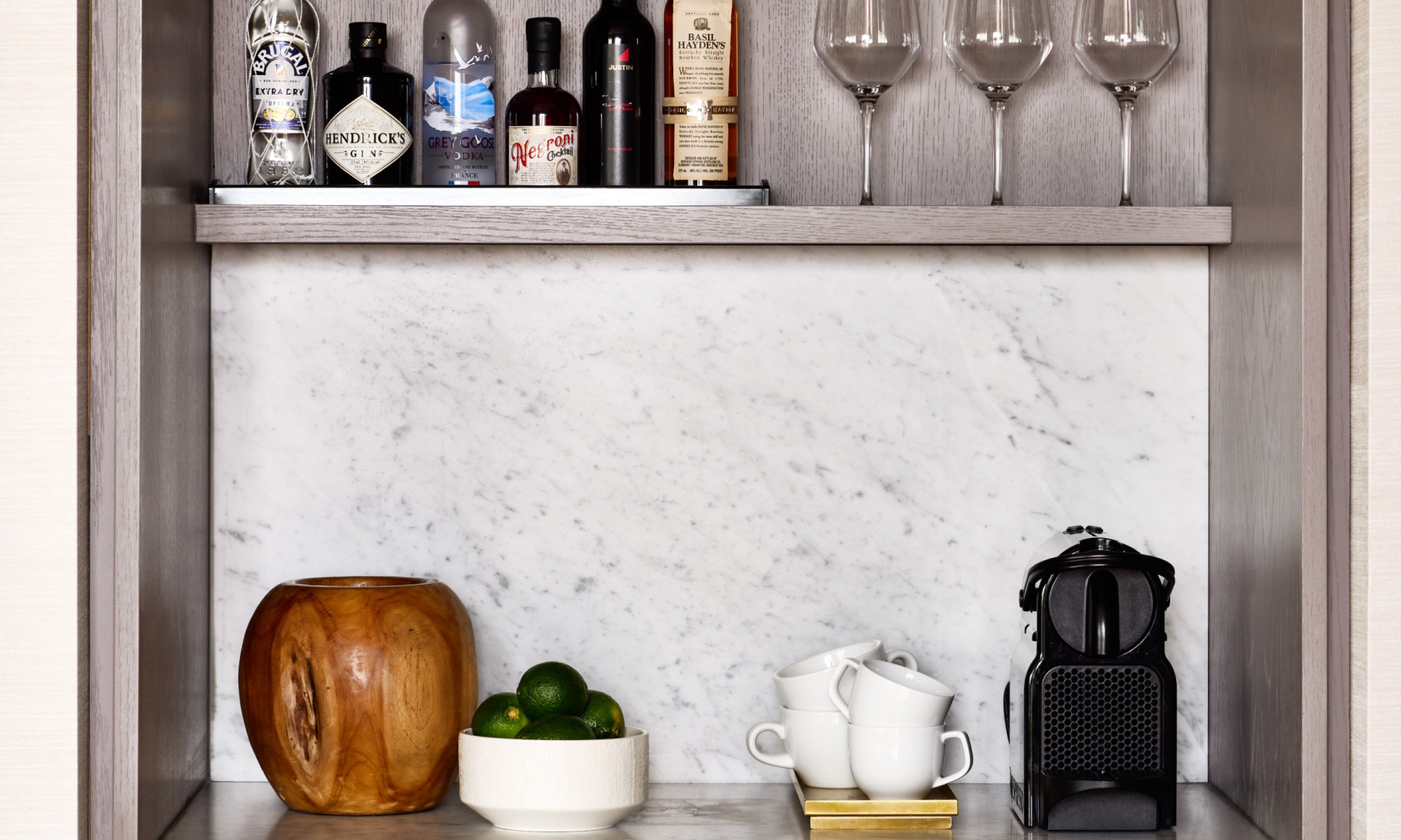 Mini bar with bottles, glasses, a small coffee maker, and a bowl of limes on a marble countertop.