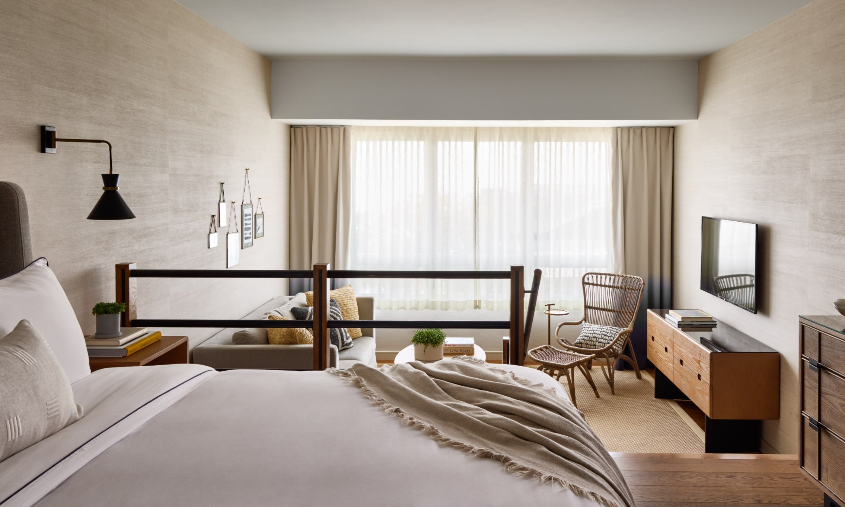 Modern hotel room with a neatly made bed, wall-mounted lamp, wood sideboard, armchair, and large window with light curtains.