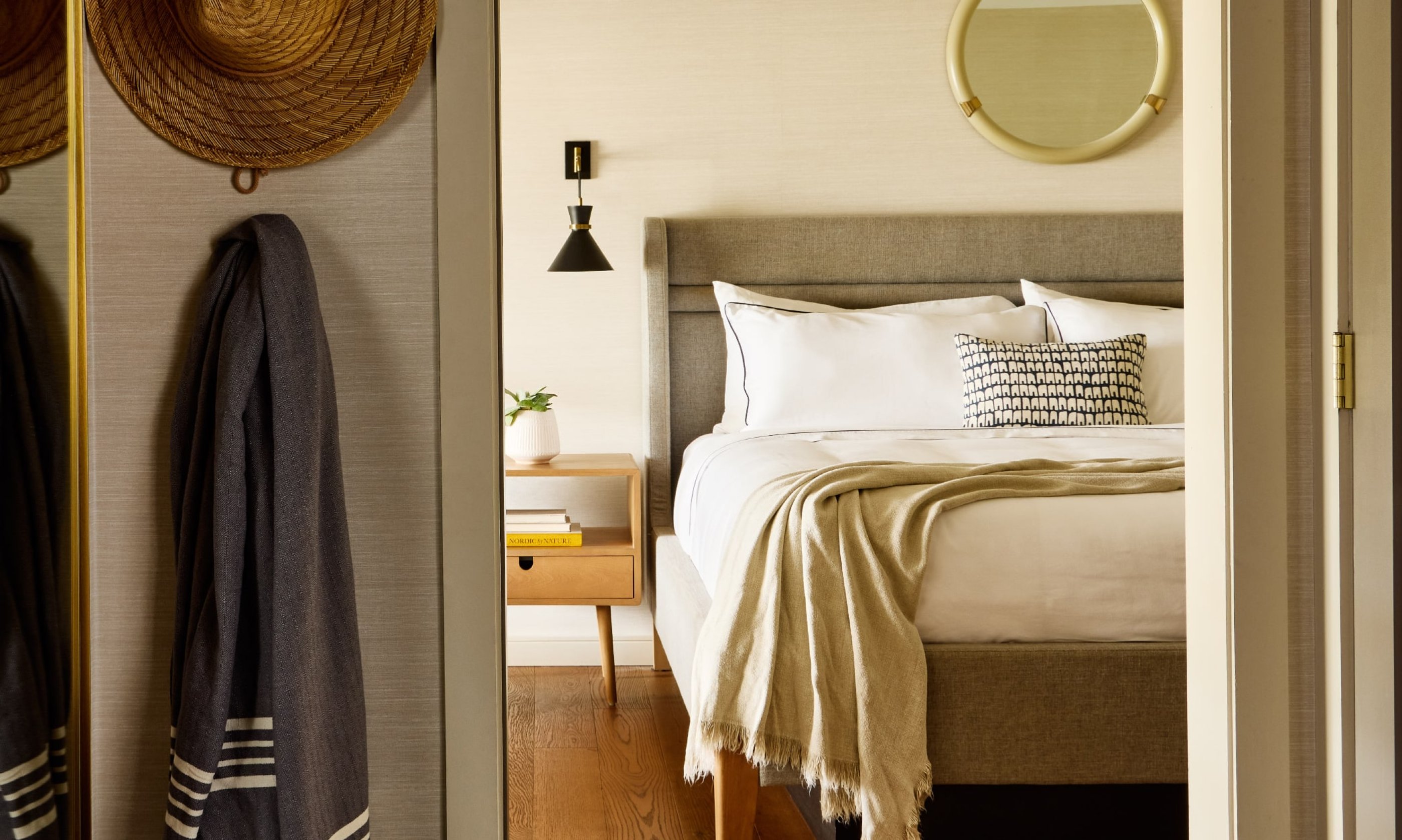 Cozy bedroom with a neatly made bed, throw blanket, and decorative pillows. A round mirror and wall lamp above the bed, with hats hanging on the wall.