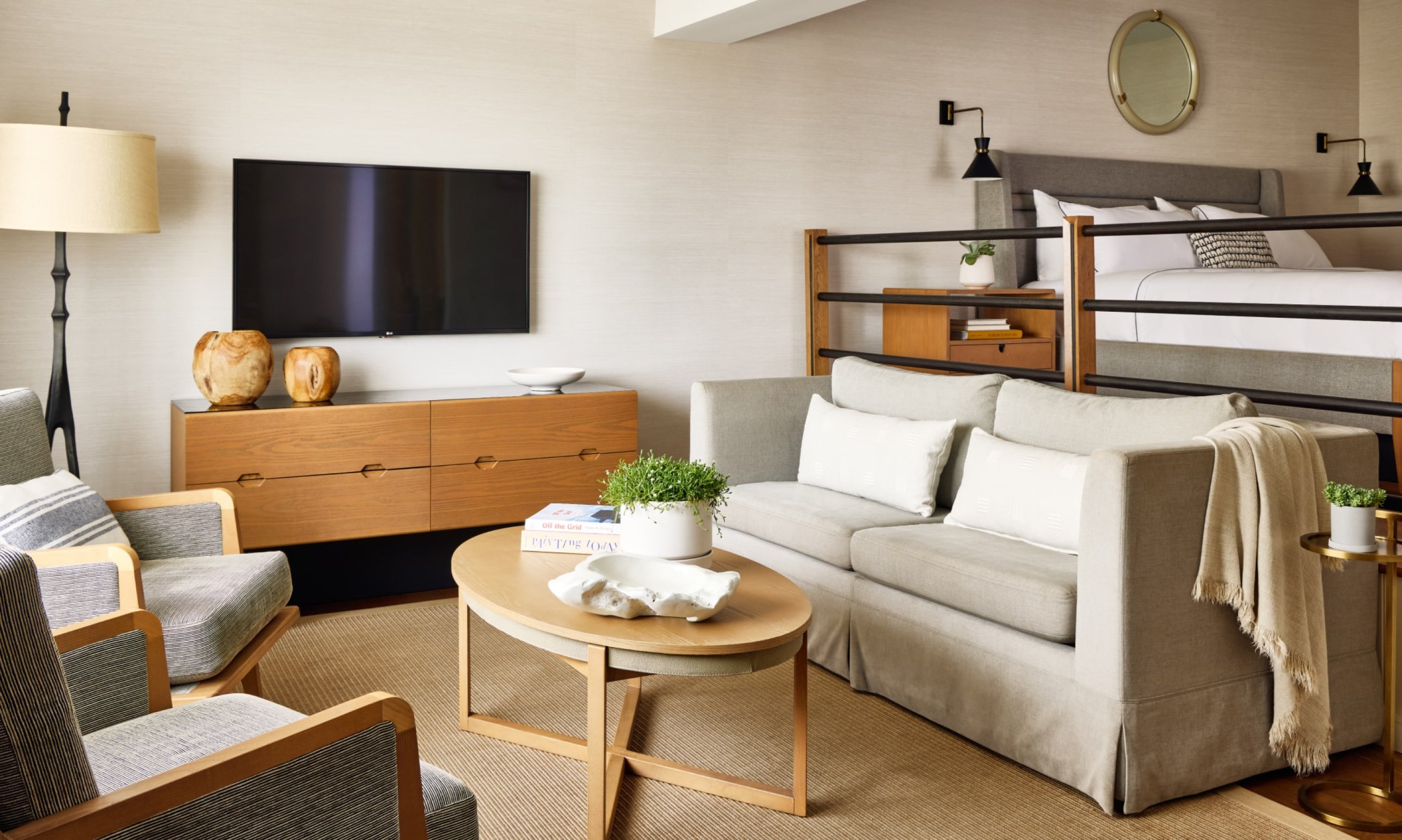 Modern living room with a beige sofa, wooden coffee table, and a wall-mounted TV. A lofted bed is visible in the background, with light neutral decor.