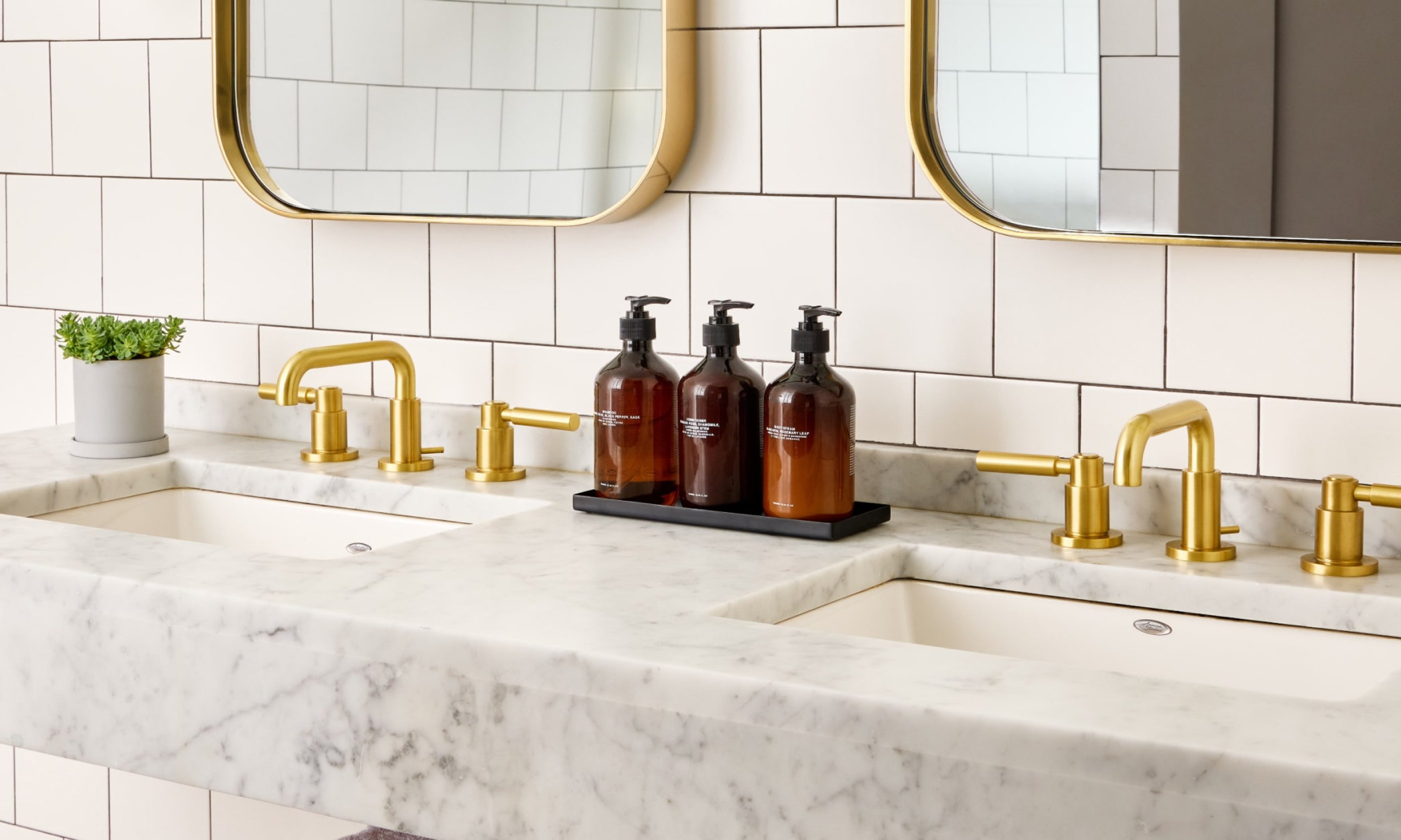 White marble bathroom countertop with two sinks, gold faucets, brown soap dispensers, and wall mirrors.