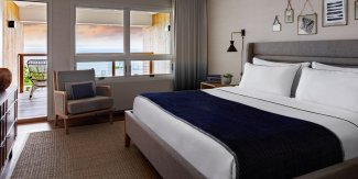 Modern hotel room with a large bed, navy blue blanket, artwork on the walls, and a wooden armchair. Window view of the ocean and sky.