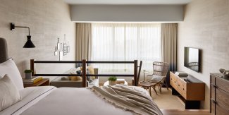 Modern hotel room with a large bed, sofa, and wall-mounted TV. Neutral colors and large window with sheer curtains create a bright, airy atmosphere.