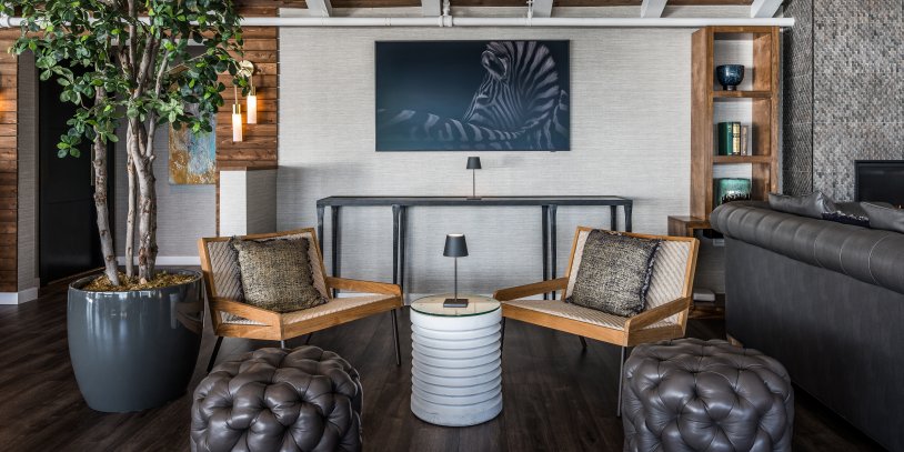 Modern living room with wooden chairs, a zebra artwork, potted tree, and tufted leather stools. Neutral tones and wood beams enhance the cozy atmosphere.