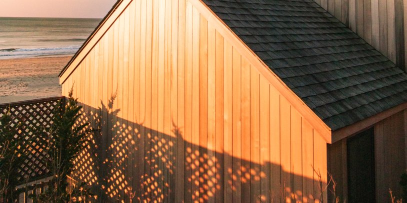 Sunlit beach house with wooden siding, casting intricate shadows on a fence. Ocean and sandy beach in the background.