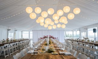 Elegant wedding reception hall with draped ceiling, round hanging lights, and long wooden tables set with white chairs and tableware.