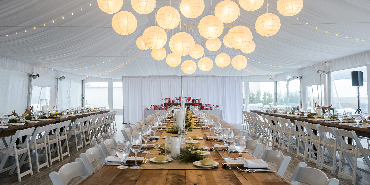 Elegant wedding reception with long tables, white chairs, and glowing hanging lanterns under a tented ceiling.