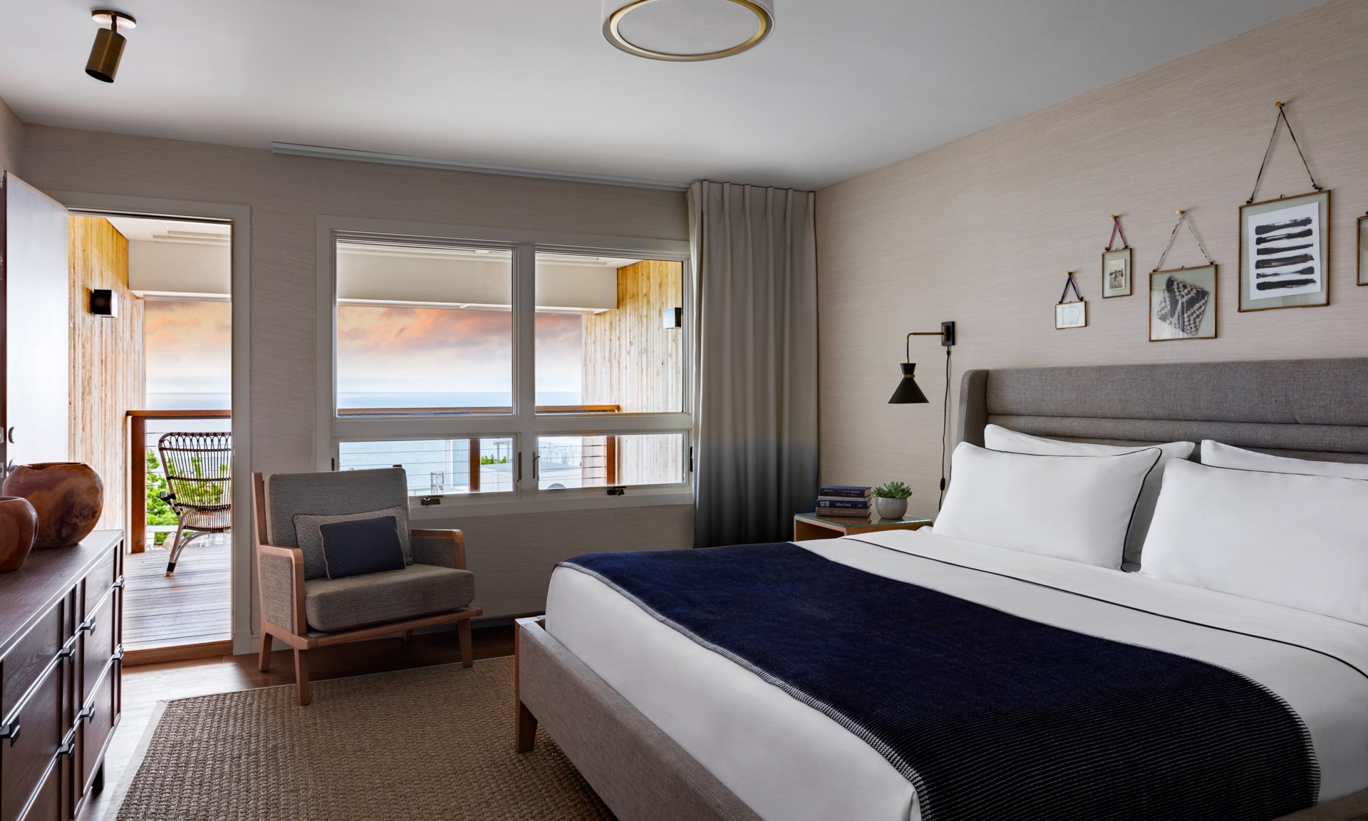 Modern hotel room with a bed, navy blanket, and pillows. Large windows show a balcony and ocean view. Minimalist decor with framed art on the wall.