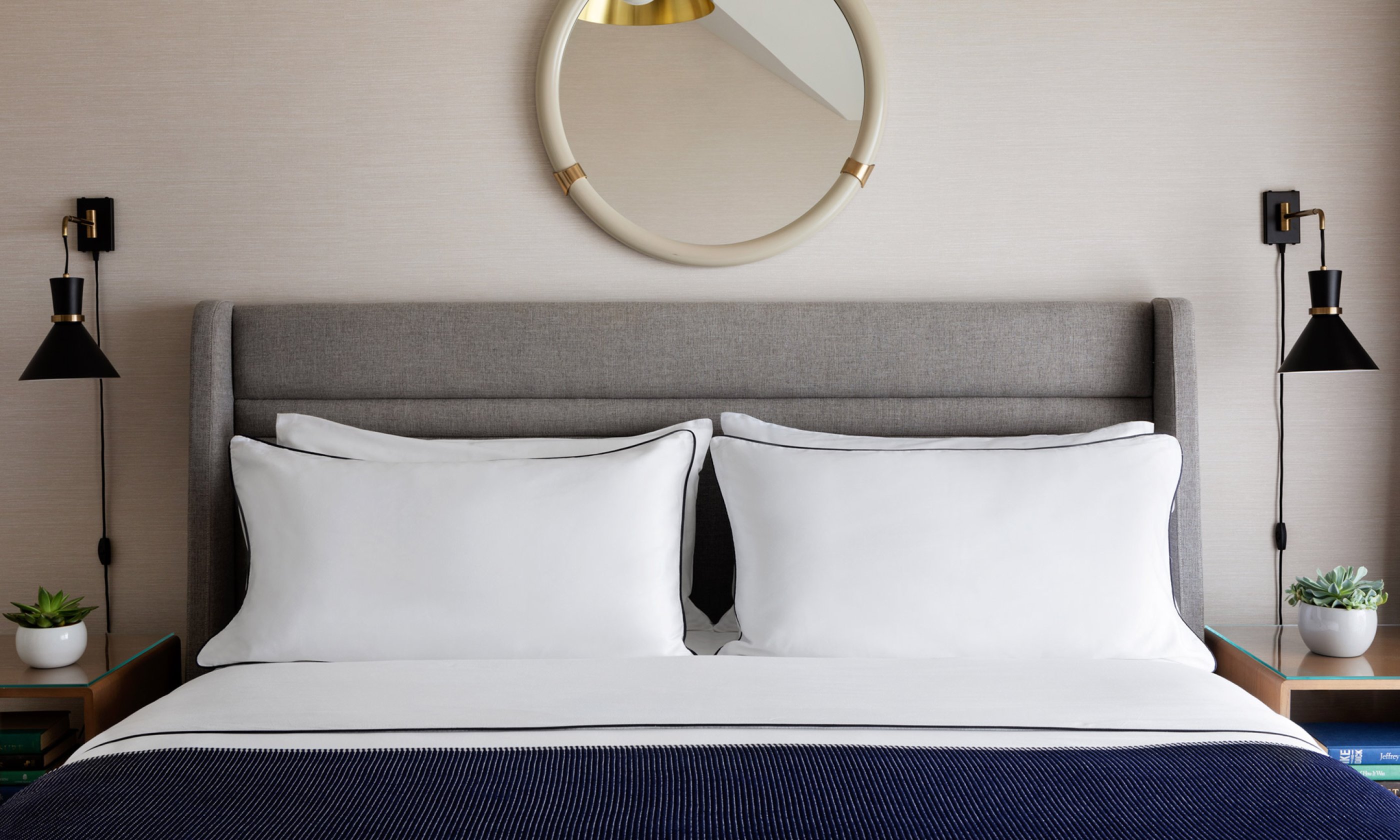 A neatly made bed with white pillows and a navy blanket, flanked by modern black lamps and small plants. A round mirror hangs above the bed.