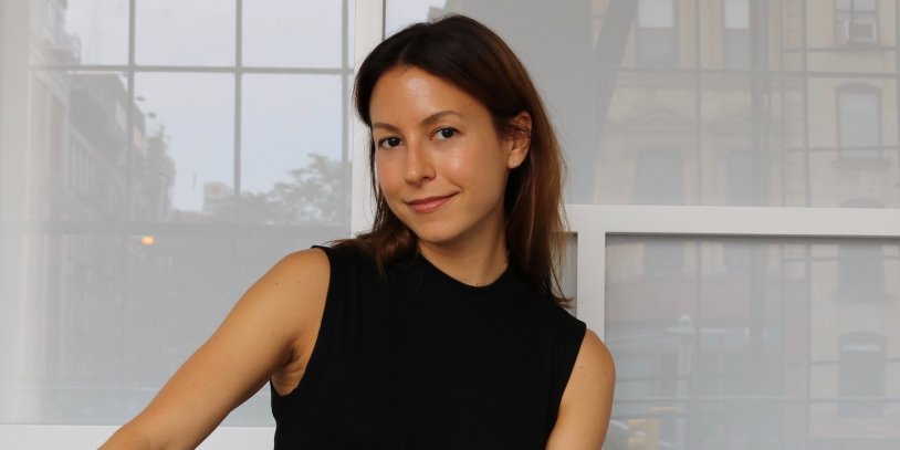 Woman with long brown hair in a sleeveless black top, smiling indoors with large windows in the background.