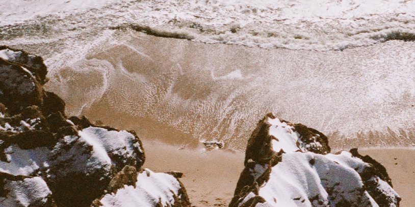Waves gently crashing on a sandy beach, flanked by snow-dusted rocks.