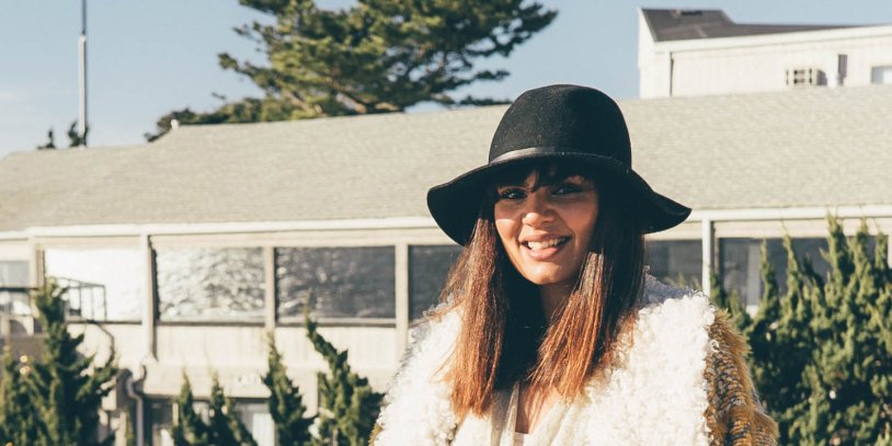 Woman wearing a black hat and a fluffy white coat, smiling outdoors with trees and a building in the background.