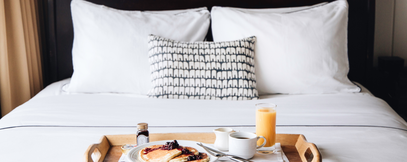 Breakfast tray with pancakes, juice, and coffee on a neatly made bed with white pillows and a patterned cushion.